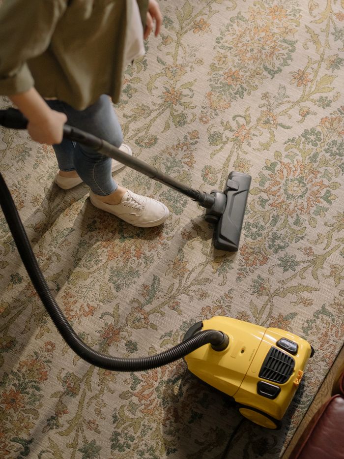 Personne nettoyant un tapis fleuri avec un aspirateur jaune à l’intérieur.